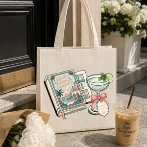 May include: A cream-colored canvas tote bag featuring a graphic print of an open book titled Salt Air & Sweet Consequences, a cocktail glass with a lime slice and a pink bow, and a small tag labeled Lathel Serenade with a seashell icon.
