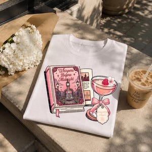May include: A white t-shirt featuring a graphic print of a pink book titled Whispers Before Midnight, an open book page, and a pink cocktail glass garnished with a raspberry and a pink bow, with a small tag labeled Intoxicating Elixir.