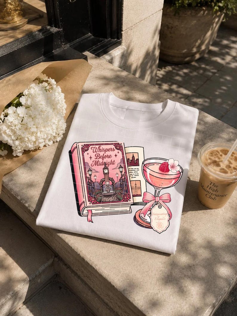 May include: A white t-shirt featuring a graphic print of a pink book titled Whispers Before Midnight, an open book page, and a pink cocktail glass garnished with a raspberry and a pink bow, with a small tag labeled Intoxicating Elixir.