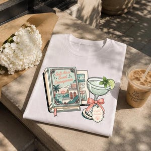 May include: A white t-shirt featuring a graphic print of an open book titled Salt Air & Sweet Consequences, depicting a coastal scene with a sunset, palm trees, and a building, alongside a cocktail glass with a lime slice and a pink bow tag.