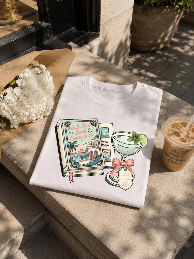 May include: A white t-shirt featuring a graphic print of an open book titled Salt Air & Sweet Consequences, depicting a coastal scene with a sunset, palm trees, and a building, alongside a cocktail glass with a lime slice and a pink bow tag.