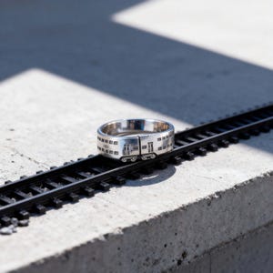 Op de afbeelding: Een zilveren ring ontworpen als een treinwagon, met ramen en deuren. Hij ligt op een miniatuur zwart treinspoor, tentoongesteld op een betonnen oppervlak. Het ontwerp van de ring is gedetailleerd.