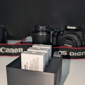 Könnte beinhalten: Drei weiße Canon LP-E8 Kamera-Akkus sind in einer schwarzen Kunststoff-Aufbewahrungsschale angeordnet. Die Akkus tragen aufgedruckten Text, einschließlich Modellname und Sicherheitshinweise. Eine schwarze Canon EOS Digitalkamera ist im Hintergrund.
