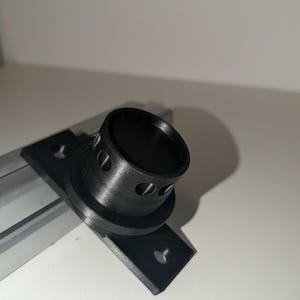 Könnte beinhalten: Eine schwarze 3D-gedruckte Montagehalterung mit einem zylindrischen Mittelteil und einer Reihe kleiner kreisförmiger Aussparungen am Rand. Die Basis ist rechteckig mit zwei vorgebohrten Löchern zur Befestigung an einer silbernen Aluminiumprofilschiene.
