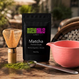 Könnte beinhalten: Ein schwarzer Beutel MatchaMallow Premium Grade Matcha-Pulver mit einem Nettogewicht von 30 g, begleitet von einem hellbraunen Bambusbesen, einem silbernen Metall-Messlöffel mit grünem Pulver und einer einfarbigen rosa Keramikschale.