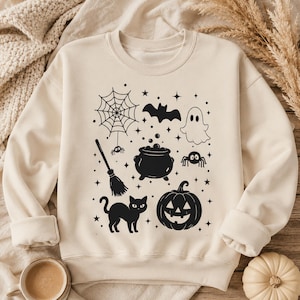 Könnte beinhalten: Ein cremefarbenes Sweatshirt mit Rundhalsausschnitt und einem schwarzen Grafikdruck von Halloween-Motiven, darunter ein Spinnennetz, eine Fledermaus, ein Geist, ein Besen, ein Kessel, eine Spinne, eine schwarze Katze, ein Kürbis und kleine Sterne.