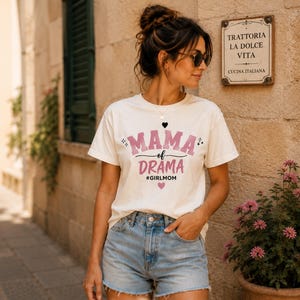Könnte beinhalten: Cremefarbenes T-Shirt mit dem Text "MAMA of DRAMA #GIRLMOM" in Rosa und Schwarz. Das Shirt wird mit Jeansshorts kombiniert. Im Hintergrund ist ein Schild für ein italienisches Restaurant zu sehen.