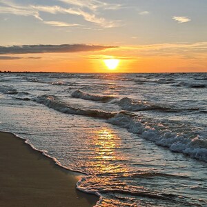 May include: A sunset over the ocean with a bright orange sun low on the horizon. Gentle waves with white foam roll onto a smooth, wet sandy beach. The sky features wispy clouds illuminated by the golden light, reflecting on the water surface.