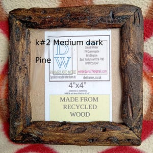 May include: A square picture frame crafted from dark, textured recycled pine wood. The frame features a label with the text k#2 Medium dark Pine, 4"x4", and MADE FROM RECYCLED WOOD, along with contact details for DW Frames and More in Bridlington.