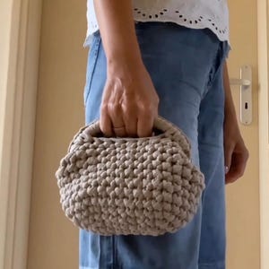 Può includere: Una borsa a mano all'uncinetto color beige con una superficie arrotondata e testurizzata e una chiusura a telaio strutturata. La borsa presenta una costruzione in filato spesso e annodato con un motivo coerente e un manico integrato.