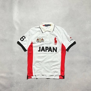 May include: A white short-sleeve polo shirt featuring red side panels and black sleeve cuffs. The front displays the word JAPAN in black, a red embroidered polo player logo, and a crest with the number 6. The right sleeve has a black number 6.
