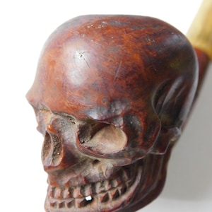 May include: A tobacco pipe with a bowl carved into the shape of a human skull. The skull is made of a dark brown, polished material with visible wood grain patterns, featuring deep-set eye sockets, a nasal cavity, and detailed teeth.