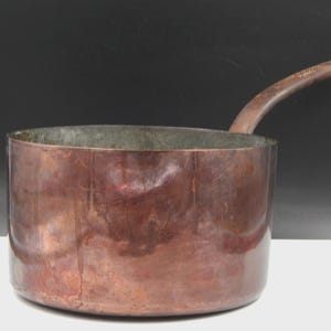 Peut inclure: Une casserole ronde en cuivre dotée d'une longue poignée incurvée s'étendant sur le côté. L'extérieur du récipient présente une finition métallique brune patinée avec des variations de surface visibles et un corps cylindrique lisse.