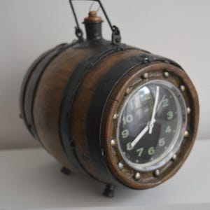 May include: A horizontal wooden barrel clock featuring dark brown wood staves held by black metal bands. The clock face is black with white numerals and hands, protected by a circular glass cover secured with metal rivets. A metal handle sits on top.