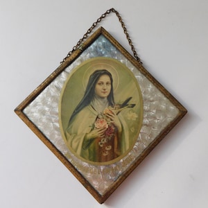 Peut inclure: Une décoration murale en forme de losange présentant un portrait ovale de Sainte Thérèse de Lisieux tenant des roses. L'image est placée sur un fond texturé, irisé et écailleux dans un cadre en bois fin avec une chaîne de suspension en métal.