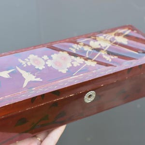 Può includere: Una scatola rettangolare in legno laccato marrone rossastro scuro con un coperchio incernierato decorato con illustrazioni dipinte color oro di due uccelli in volo e fiori. Il pannello frontale presenta una toppa circolare in metallo.
