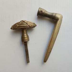 Könnte beinhalten: Ein Türknauf aus Messing mit einem strukturierten, gerippten, ovalen Kopf und einem quadratischen Metallstift, neben einem L-förmigen Türgriff aus Messing mit zylindrischem Befestigungspunkt und einem glatten, länglichen Griffbereich.