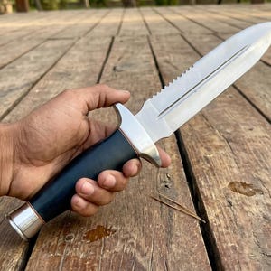 May include: A stainless steel knife with a serrated spine and a black handle. The knife has a silver bolster and a silver end cap. The knife is held against a wooden surface.