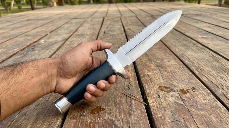 May include: A stainless steel knife with a serrated spine and a black handle. The knife has a silver bolster and a silver end cap. The knife is held against a wooden surface.