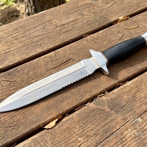 May include: A silver and black dagger with a serrated edge and a black handle. The dagger is lying on a weathered wooden surface, showcasing its sharp design and craftsmanship. The blade is pointed and the handle has a silver metal cap.