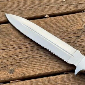 May include: A stainless steel knife with a serrated edge and a black handle. The blade is sharp and pointed. The knife is lying on a wooden surface.