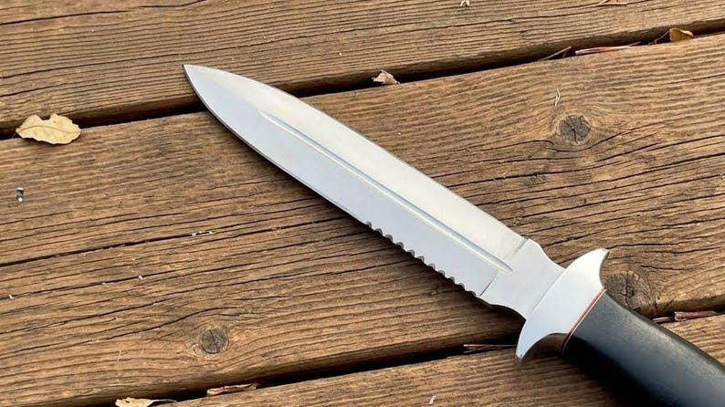 May include: A stainless steel knife with a serrated edge and a black handle. The blade is sharp and pointed. The knife is lying on a wooden surface.