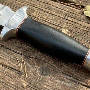 May include: A close-up of a survival knife with a serrated blade and a black handle. The knife has a silver bolster and a copper-colored accent. The knife is lying on a wooden surface.