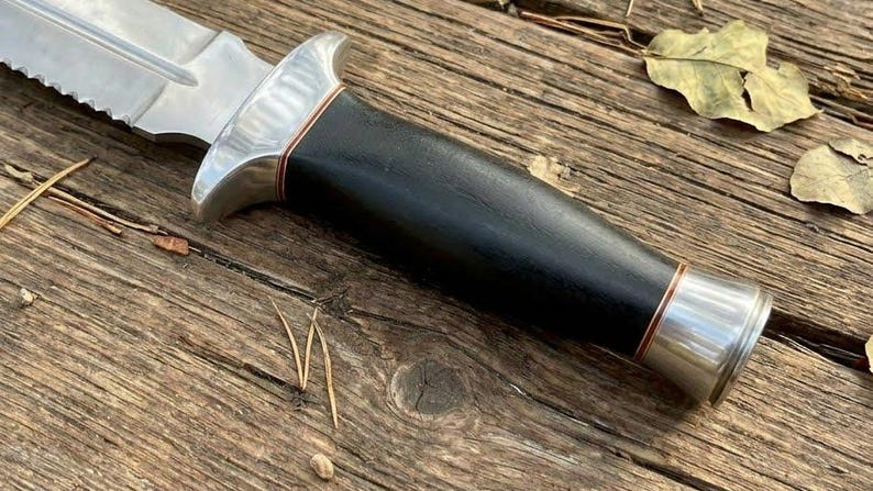 May include: A close-up of a survival knife with a serrated blade and a black handle. The knife has a silver bolster and a copper-colored accent. The knife is lying on a wooden surface.