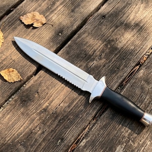 May include: A silver and black dagger with a serrated edge and a black handle. The knife is lying on a weathered wooden surface with a few fallen leaves.