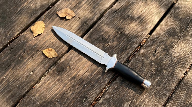 May include: A silver and black dagger with a serrated edge and a black handle. The knife is lying on a weathered wooden surface with a few fallen leaves.