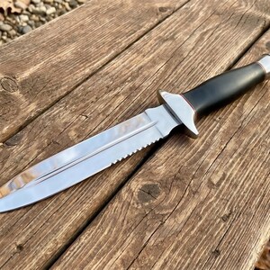 May include: A stainless steel knife with a serrated edge and a black handle. The knife has a silver bolster and a pointed blade. The knife is lying on a wooden surface.