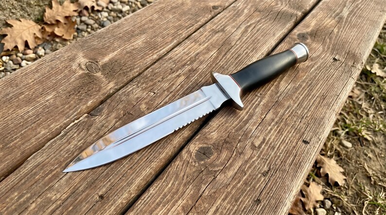 May include: A stainless steel knife with a serrated edge and a black handle. The knife has a silver bolster and a pointed blade. The knife is lying on a wooden surface.