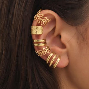 Peut inclure: Un ensemble de sept bagues d'oreille dorées portées le long du bord externe de l'oreille. La collection comprend deux bagues ajourées à motif de feuilles, une large bague texturée, deux anneaux lisses polis et deux anneaux plus fins détaillés.