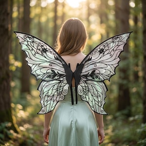 Puede incluir: Grandes alas de estilo mariposa iridiscente con intrincados patrones de filigrana negra y una estructura central negra. Las alas se llevan sobre un vestido sin mangas de color verde claro, mostrando una superficie translúcida y brillante con líneas detalladas.