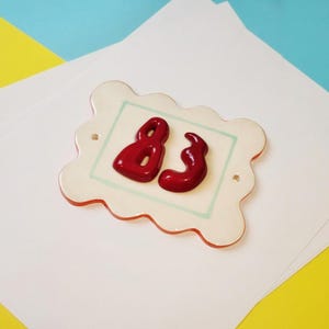 May include: A rectangular ceramic house number plaque with a scalloped edge, featuring a glossy white finish, a thin light green border, and the number 83 in raised, dark red, rounded, glossy lettering, with two small holes for mounting.