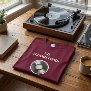 Peut inclure: Un t-shirt bordeaux avec les mots "NO ALGORITHMS" et un graphique de disque vinyle. Une platine tourne-disque avec un disque vinyle en rotation est en arrière-plan, ainsi qu'une tasse de café et une pile de disques. La scène se déroule sur une table en bois.
