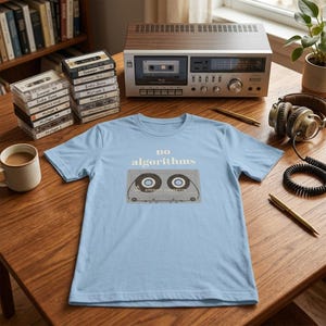 Peut inclure: Un t-shirt bleu clair avec l'inscription "no algorithms" et un dessin de cassette audio. Le t-shirt est posé sur une table en bois avec une chaîne stéréo vintage, des écouteurs et des cassettes.