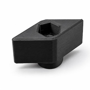 Könnte beinhalten: Ein schwarzes, geometrisch geformtes Objekt mit einer sechseckigen Aussparung. Das Objekt hat einen zylindrischen Sockel und eine dreieckige Oberseite mit einer sechseckigen Öffnung. Die Oberfläche ist strukturiert.