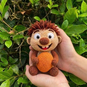 Può includere: Bambola all'uncinetto fatta a mano con un viso sorridente, capelli castani e grandi occhi. La bambola ha braccia e gambe marroni e tiene in mano una palla arancione. La bambola è tenuta in due mani su uno sfondo di foglie verdi.