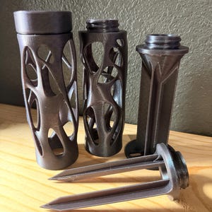 May include: A collection of four dark gray, 3D-printed objects. Two cylindrical components feature an intricate, organic web-like cutout pattern. One component is a vertical post with a circular base, and one is a pointed stake with two prongs.