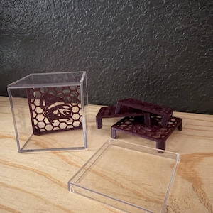 May include: A clear acrylic cube container with a removable lid, containing a dark purple, square, honeycomb-patterned platform. Beside the cube are two additional dark purple, square, honeycomb-patterned platforms with small legs.