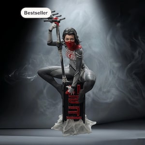 May include: A statue of a female superhero in a gray and white suit, red mask, and scarf, holding a stylized weapon. The figure is kneeling on a black platform with red lettering, atop a white web-like base. The word "Bestseller" is in a white oval.