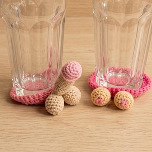 May include: Two clear glass tumblers with vertical fluted sides, each resting on a round, bright pink crocheted coaster. One coaster features a beige and pink phallic-shaped crochet attachment, while the other features two small beige and pink spheres.