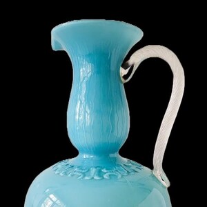 Op de afbeelding: Een lichtblauwe glazen kan met een helder, gebogen handvat. De kan heeft een bolvormige basis met een decoratief patroon en een gestructureerd oppervlak. Het handvat is bevestigd aan de bovenkant van de kan.