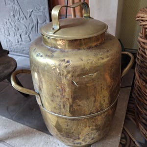 Pode incluir: Um recipiente de latão vintage com tampa e duas alças laterais. O recipiente tem formato cilíndrico com uma pátina ligeiramente envelhecida. A tampa tem uma única alça. O recipiente está sobre uma superfície de pedra.