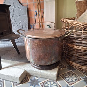 Puede incluir: Una olla grande de cobre antiguo con tapa y dos asas laterales. La olla se asienta sobre una base de madera sobre un bloque de hormigón. El cobre tiene una pátina desgastada. Una cesta de mimbre y una chimenea están en el fondo.