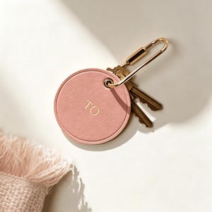 Può includere: Un portachiavi rotondo in pelle rosa con le lettere dorate "TO" e un fermaglio in metallo color oro. Diverse chiavi sono attaccate al portachiavi. Il portachiavi è su una superficie bianca.