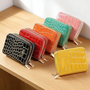 Puede incluir: Una colección de seis carteras rectangulares con una textura brillante de relieve de cocodrilo y cremalleras dobles doradas. Las carteras están disponibles en negro, rojo, naranja, verde, rosa y amarillo, algunas con nombres como Elona y Dylan.