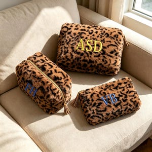 May include: Three plush leopard print pouches with gold-tone zippers and tassel pulls. One rectangular pouch features yellow embroidered letters ASD, while two smaller pouches feature blue embroidered letters FH and VB respectively.