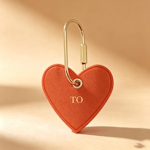 Può includere: Un portachiavi a forma di cuore in pelle arancione con chiusura color oro. Il portachiavi ha la scritta "TO" stampata sul davanti. La chiusura è in metallo color oro. Lo sfondo è di colore arancione chiaro.